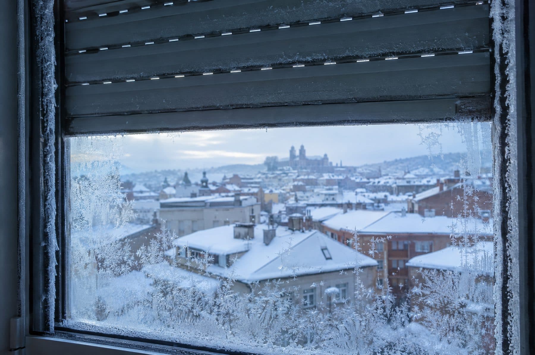 Cristal escarchado de una ventana con la persiana gris a medio bajar y tejados nevados al fondo